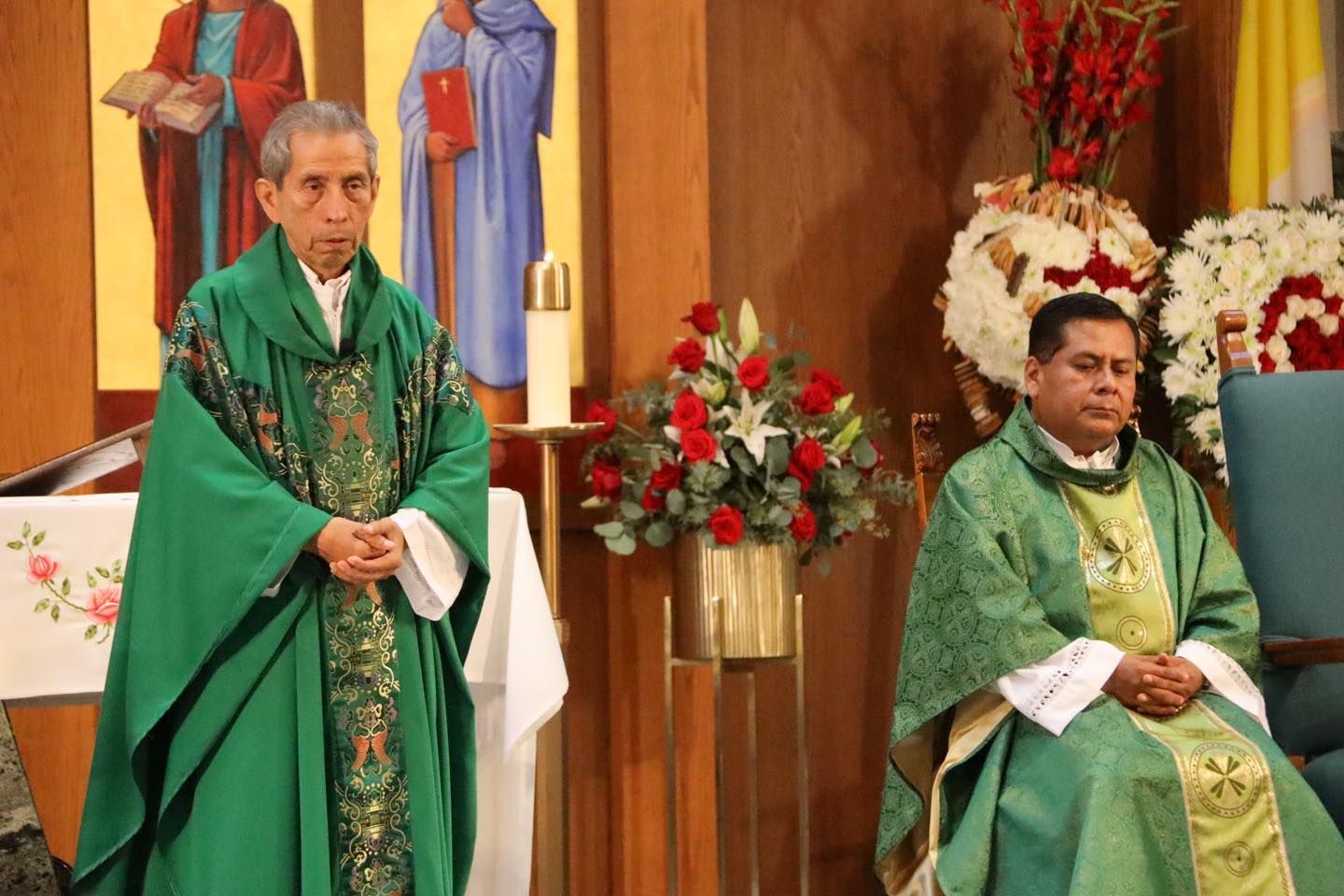 Two clergy members in green vestments stand and sit at an altar decorated with flowers.