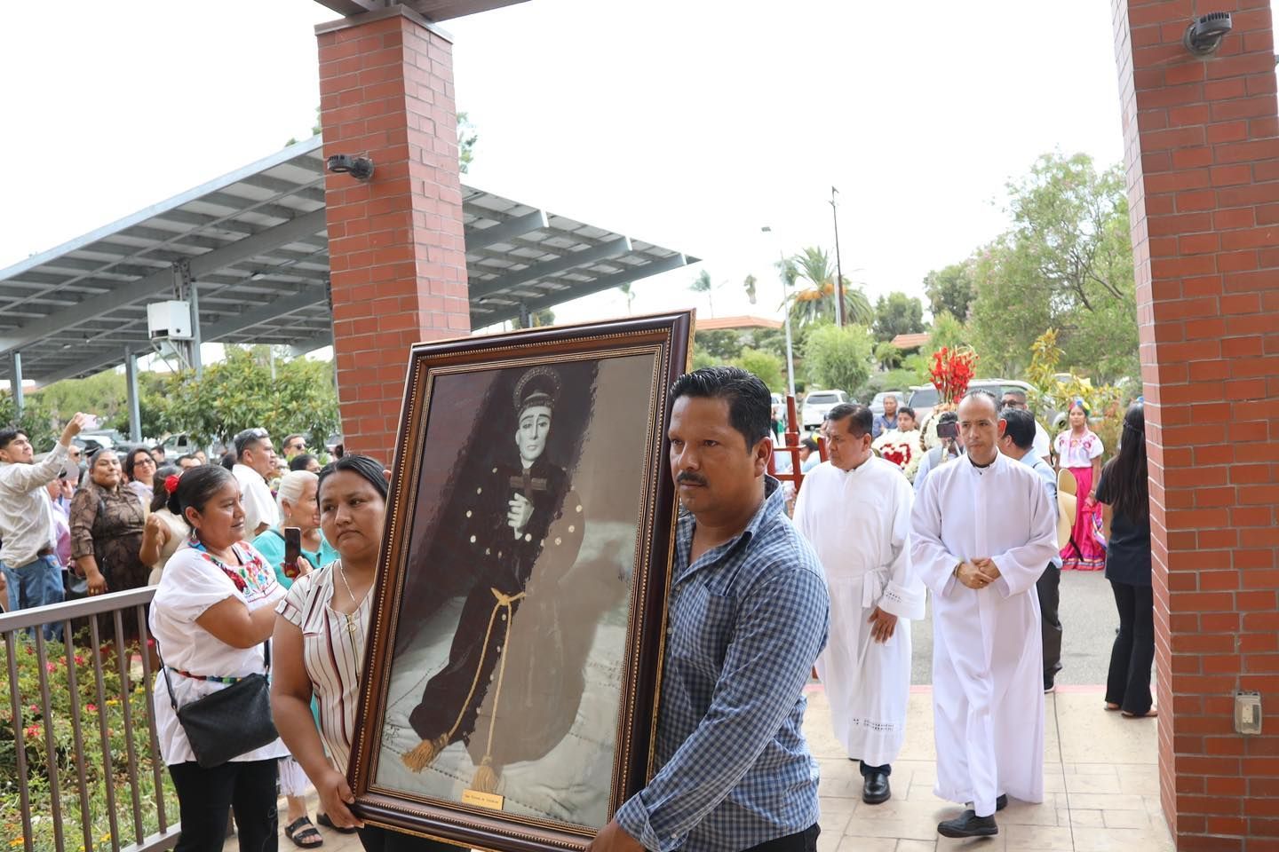 People carrying a framed portrait during a solemn outdoor procession by a brick pavilion