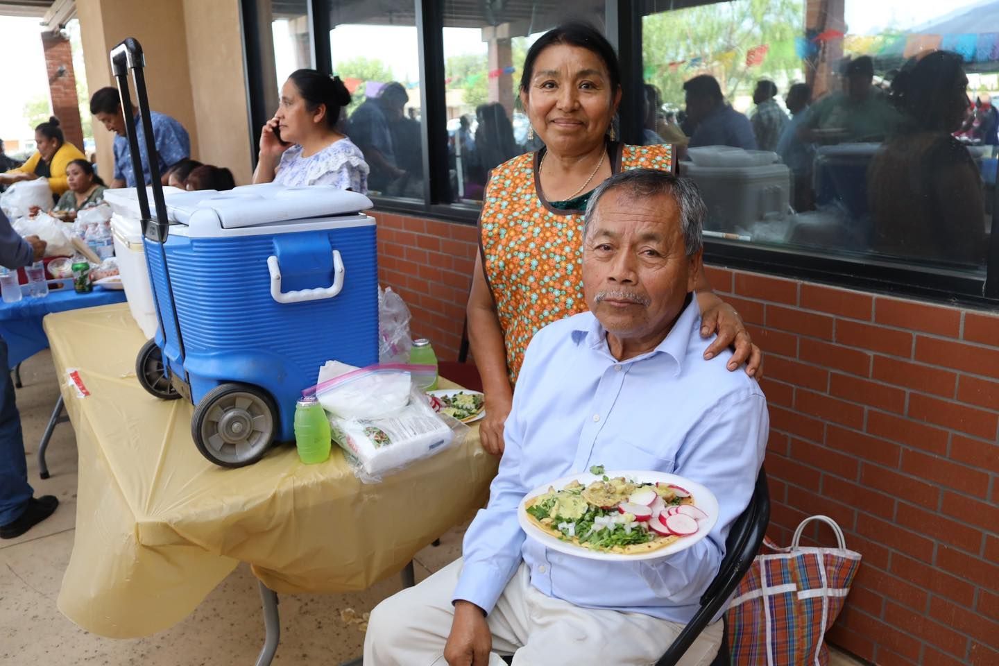 Man holding a plate of food beside a blue cooler and table at an outdoor gathering