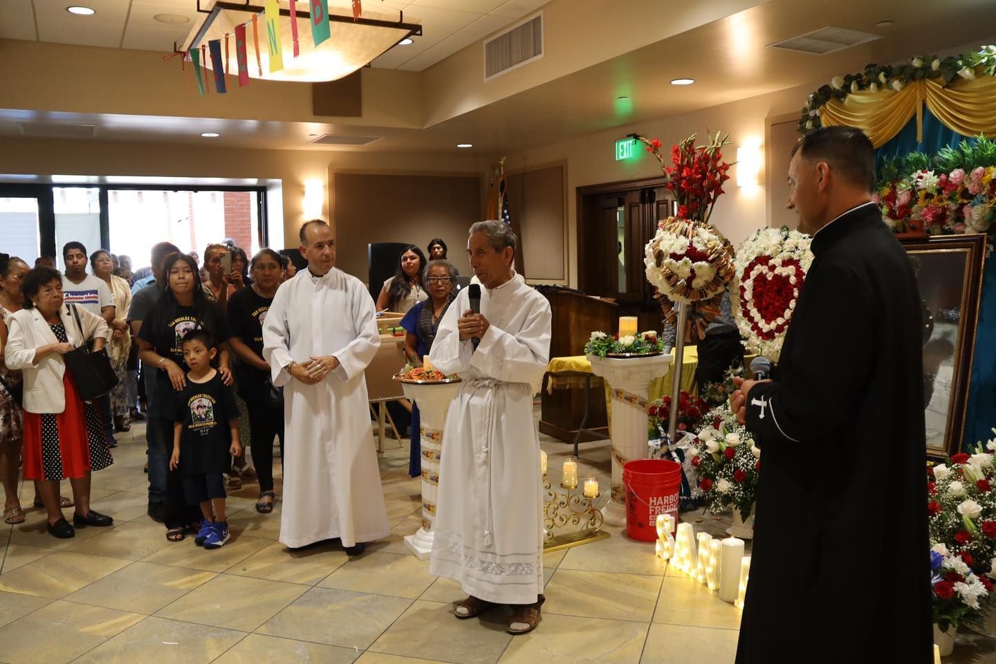 Indoor religious ceremony with people gathered around a priest at an altar decorated with flowers and candles