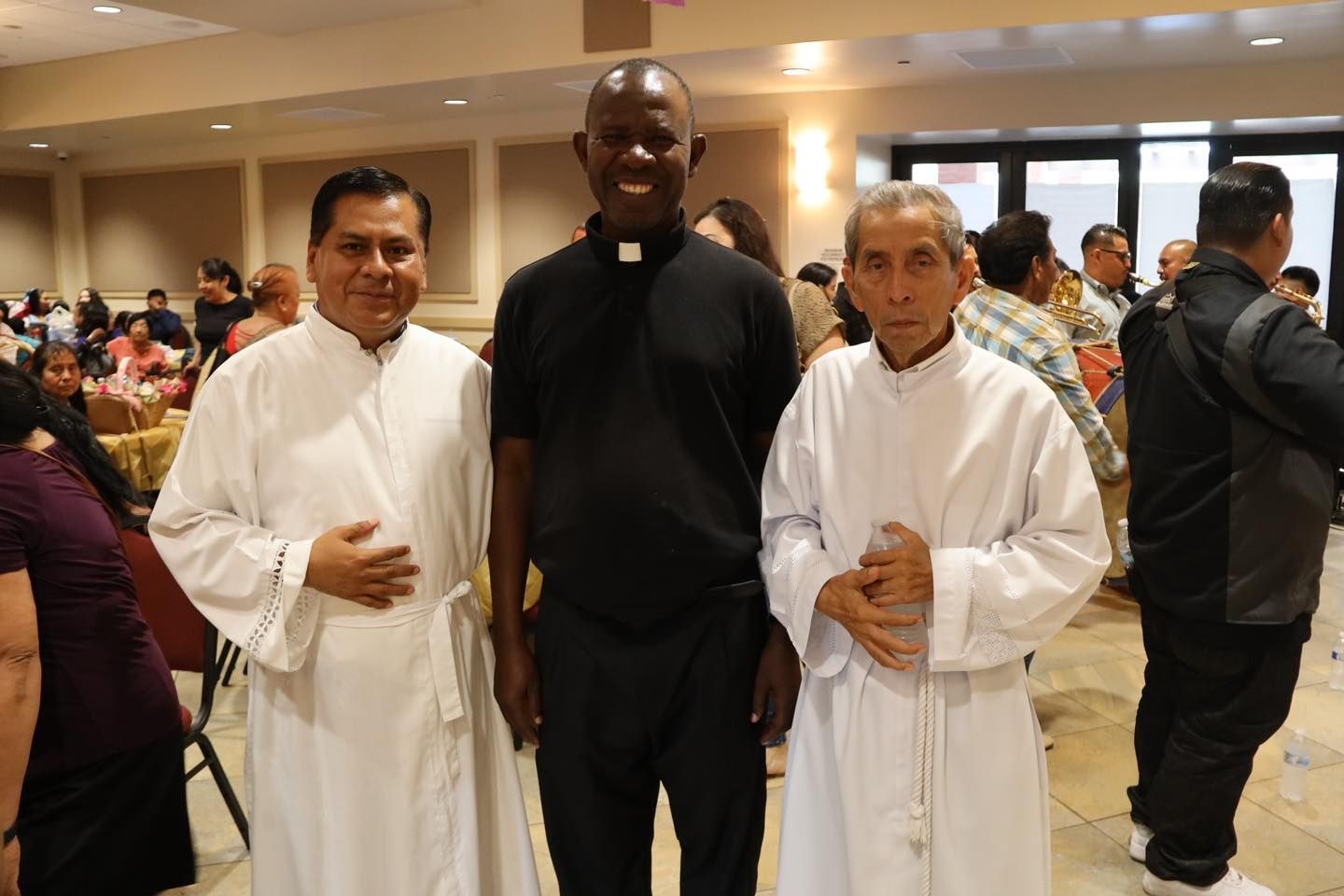 Three men in white robes stand indoors at a crowded event, posing for a photo.