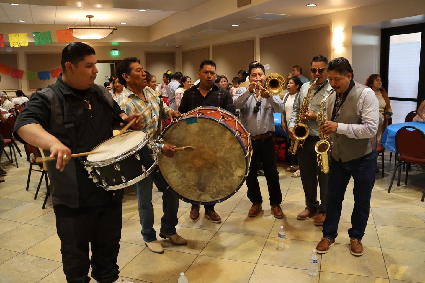 Musicians playing drums and saxophones in a hotel lobby, with a large bass drum at center.
