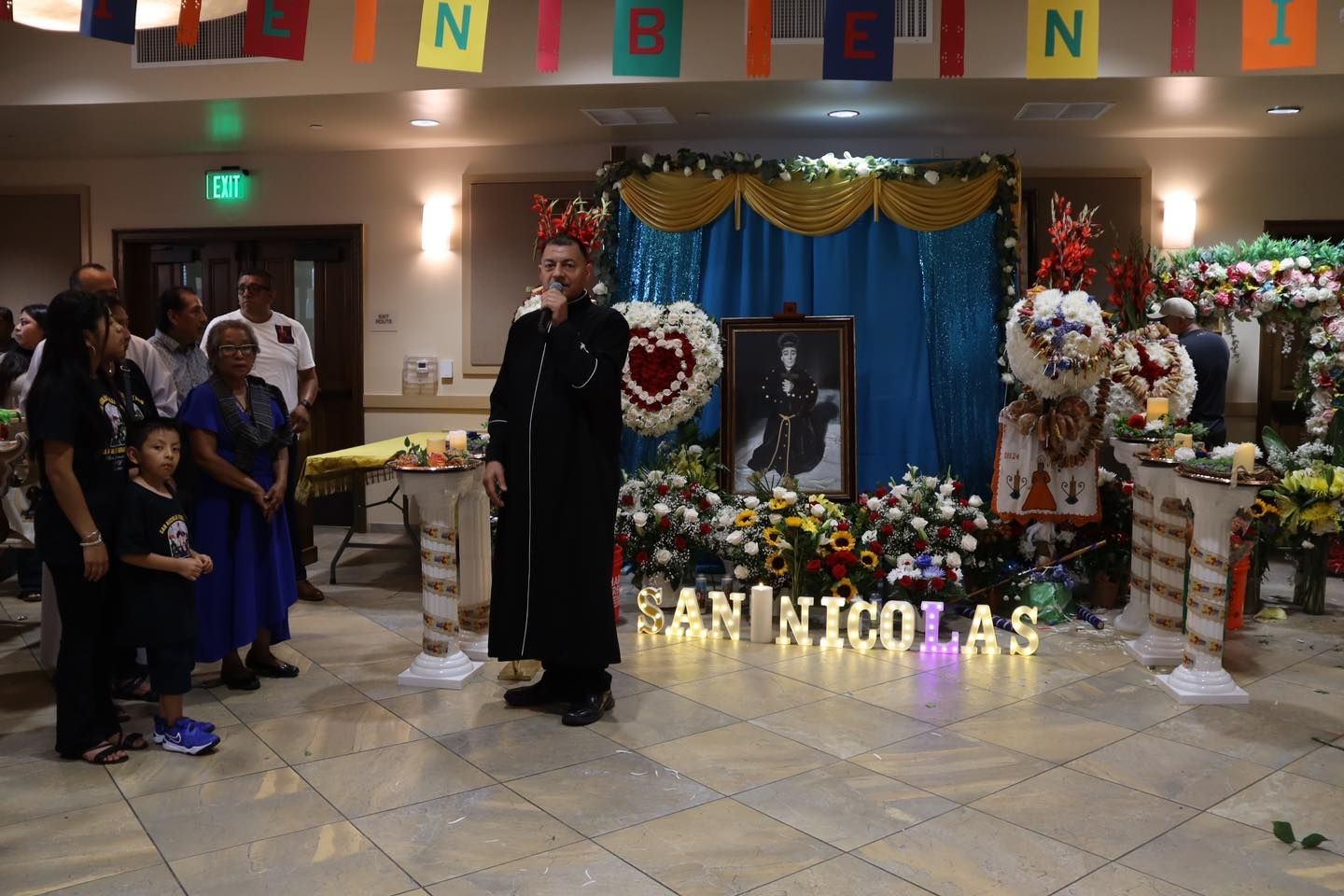People gathered in a decorated hall at a memorial display with flowers, candles, and the name “ANNICOLAS.”