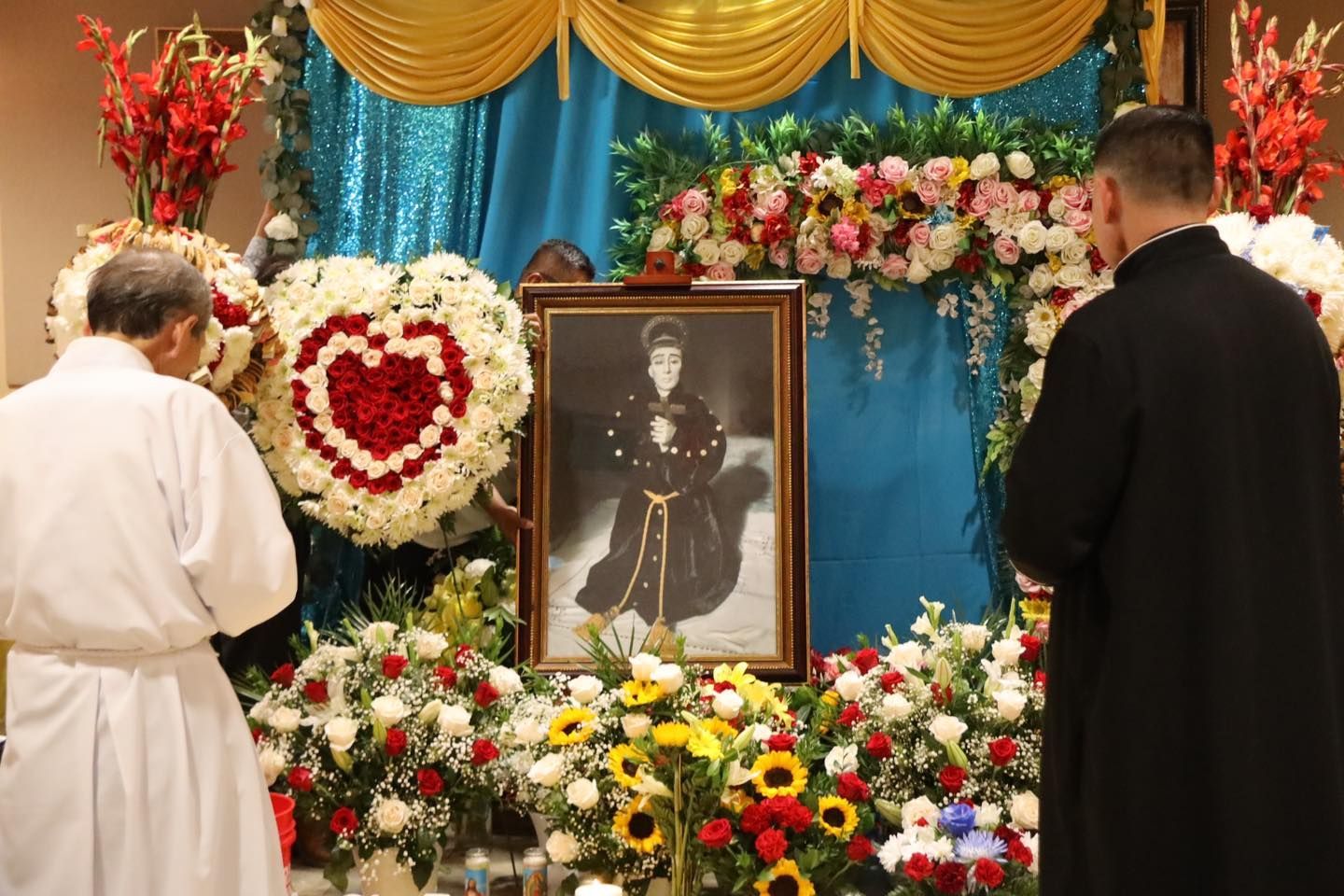 Memorial altar with framed portrait, white and red flowers, and two people standing in prayer
