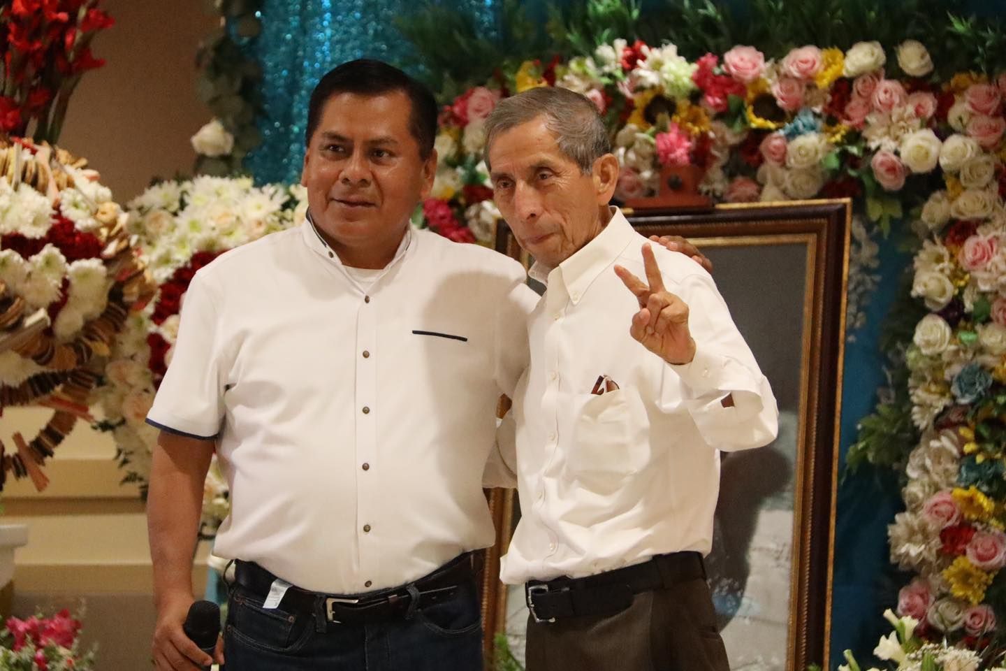 Two men in white shirts pose at a floral event backdrop, one holding up a peace sign.