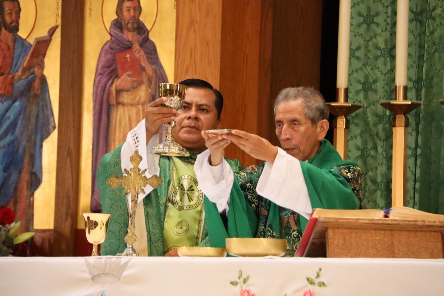 Two priests in green vestments celebrate Mass at the altar, one raising a chalice.