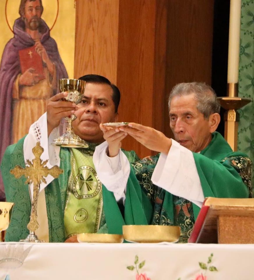 Priests in green vestments elevating a chalice and host during Mass at the altar