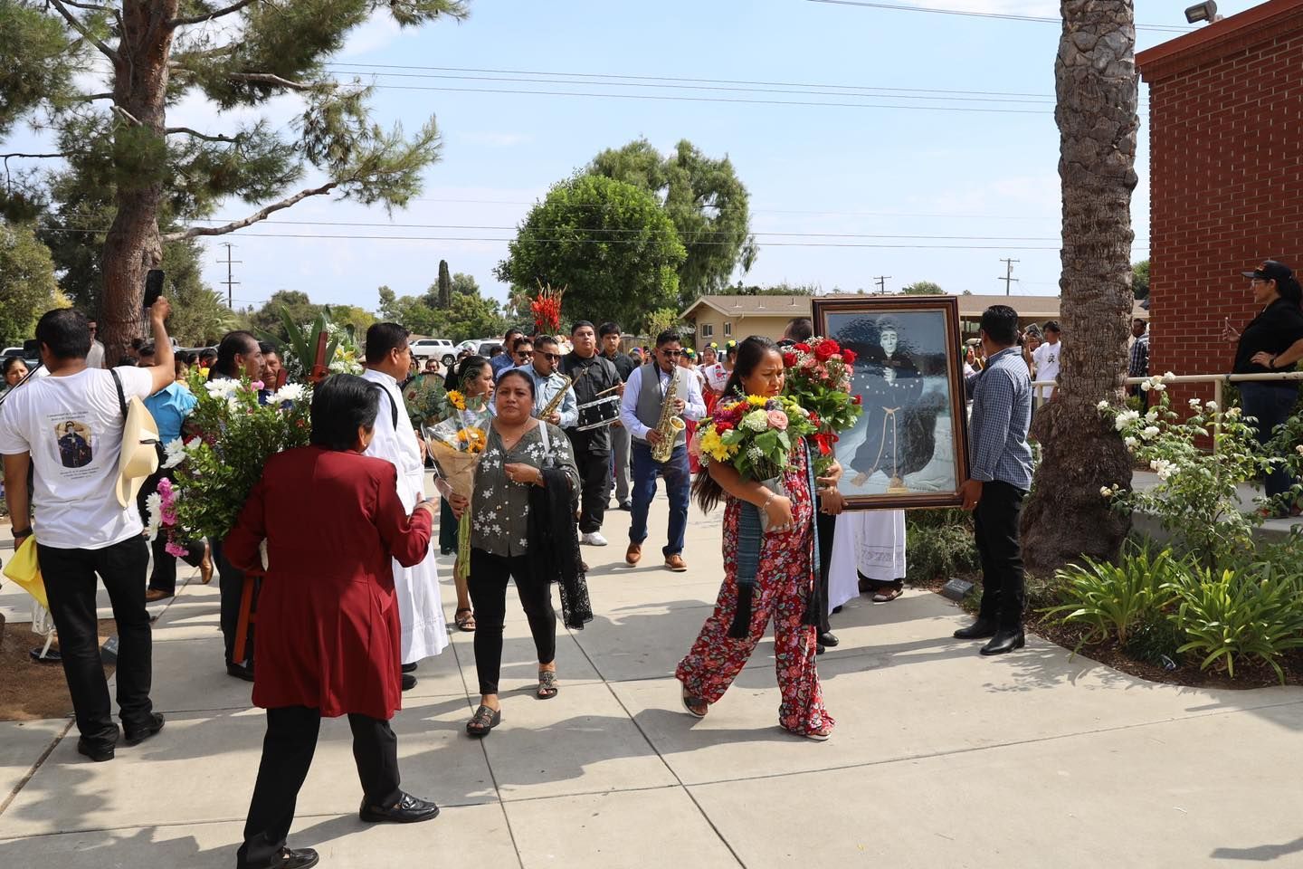 People walking outdoors on a sunny campus path, some carrying flowers and a framed portrait.