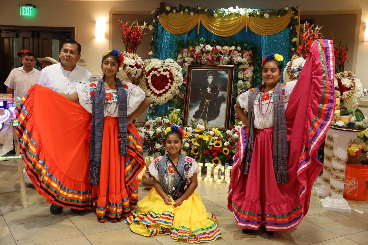 Three dancers in colorful traditional dress posing before a decorated altar and framed portrait