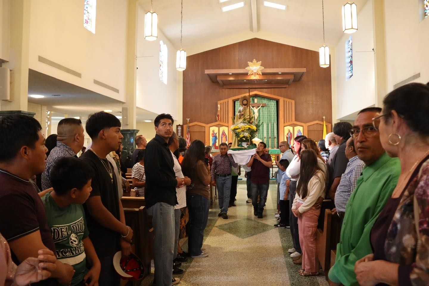 Congregation standing in church aisle during a service, facing the altar with a statue and green backdrop.
