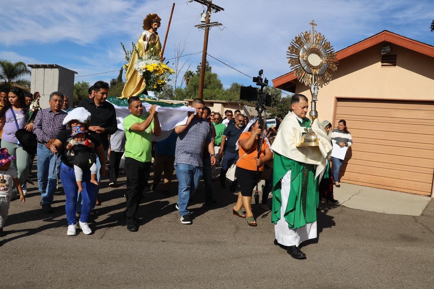 Parade procession with people carrying a statue and religious banner down a street in bright sunlight