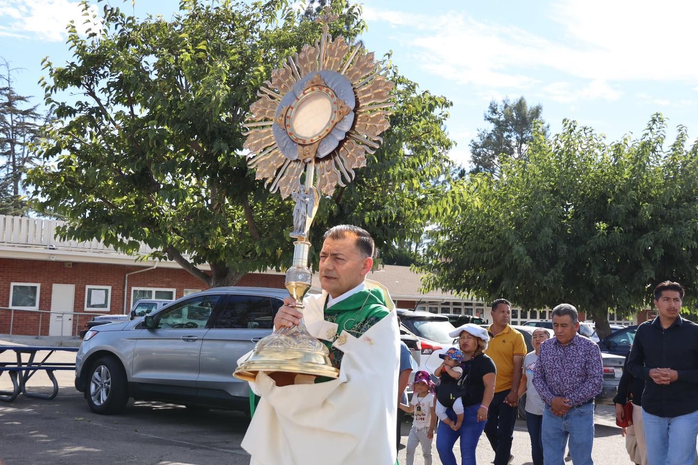 Man in white robe carrying a gold religious procession monstrance outdoors with a small group behind him