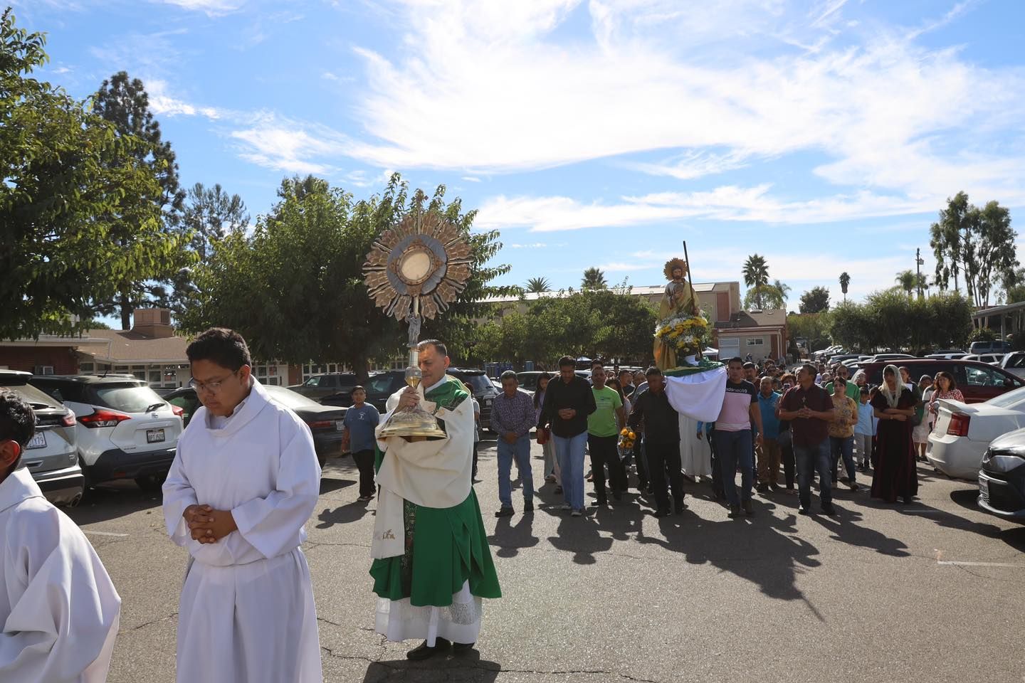 Religious procession with a priest holding a monstrance, followed by a crowd in a parking lot under a blue sky