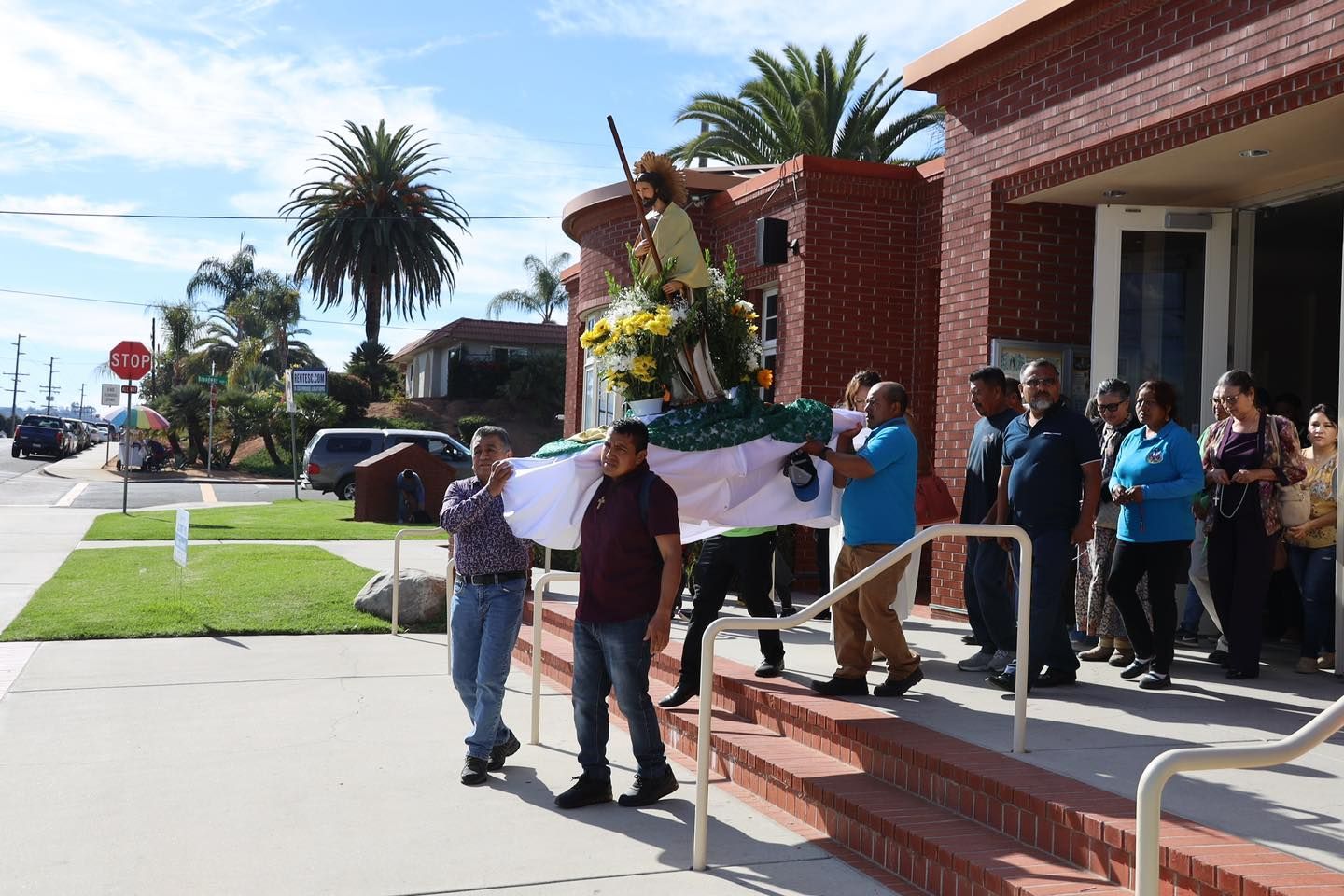 People carrying a white casket past a building, with onlookers gathered nearby.