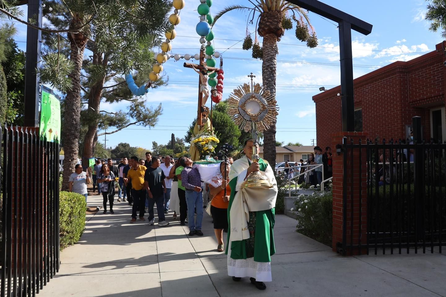 Procession with a person in white and green robes holding a decorated staff outside a red brick building