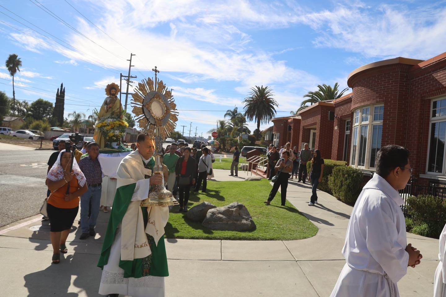 Outdoor religious procession with people carrying a decorated golden object near a church building.