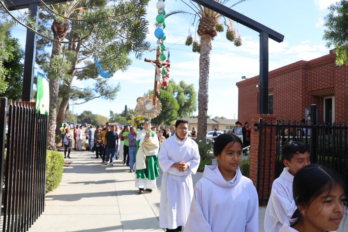 People walking in a sunny outdoor procession along a sidewalk beside a red brick building and iron fence.