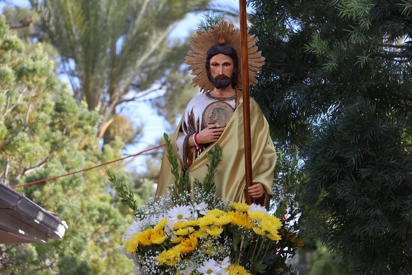 Statue of a saint in a leafy outdoor setting, holding flowers and topped with a halo