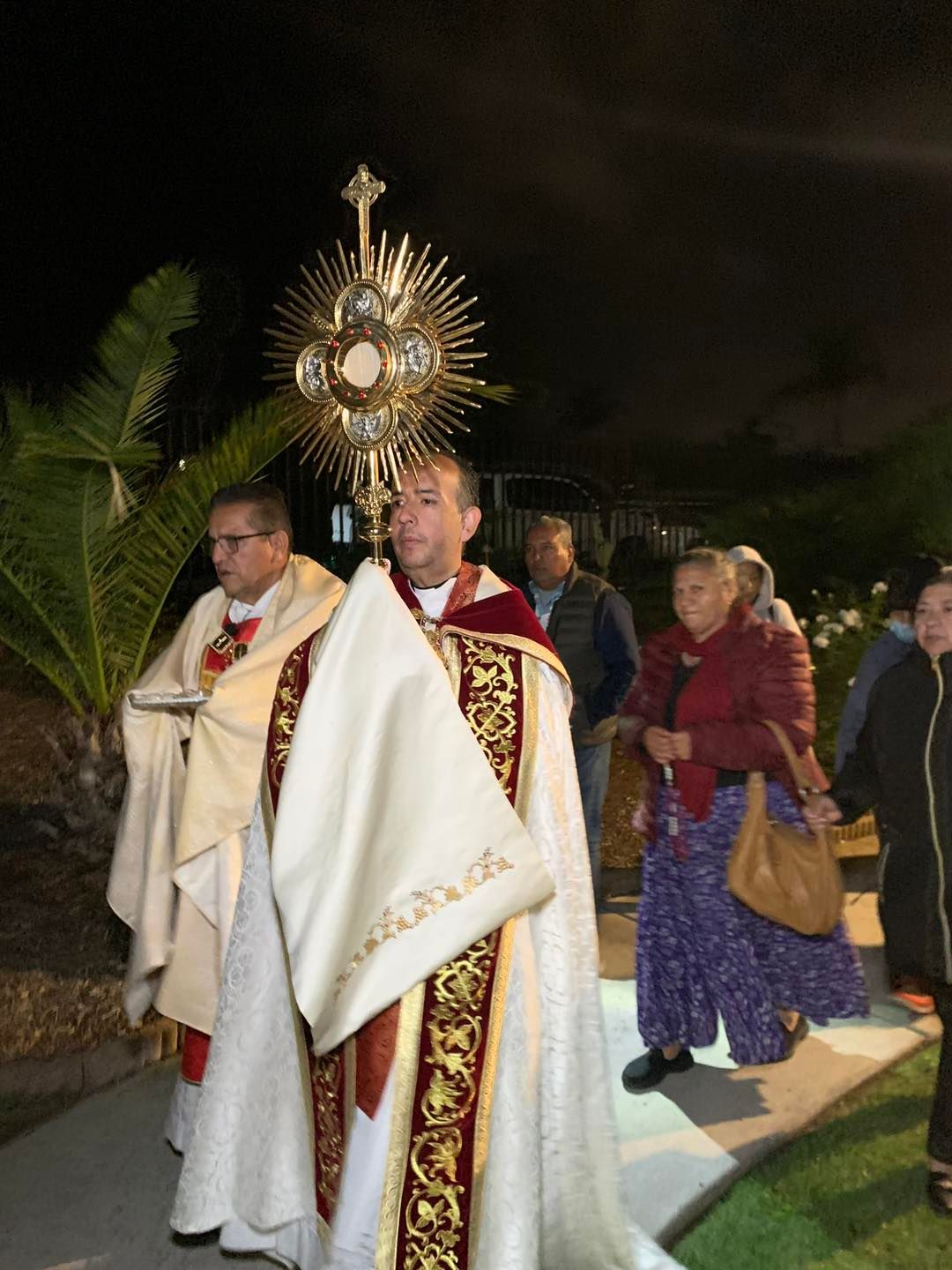 Priests in white vestments carry a golden monstrance in a nighttime outdoor procession