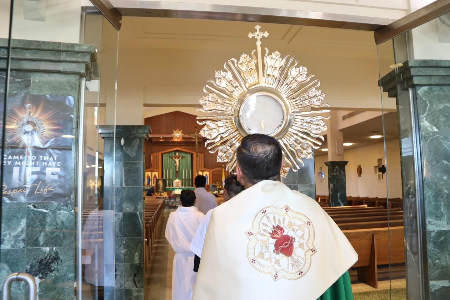 Priest in white vestments facing a large ornate monstrance inside a church sanctuary