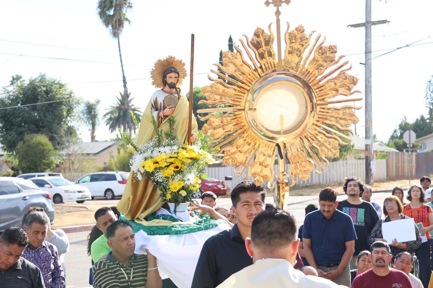 Religious procession outdoors with a saint statue and golden sunburst display, people gathered around