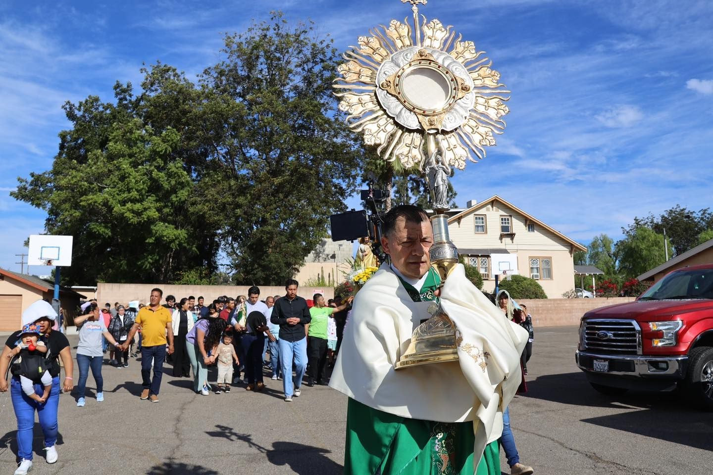 Woman in white robes carries a gold religious monstrance during a street procession under a blue sky.