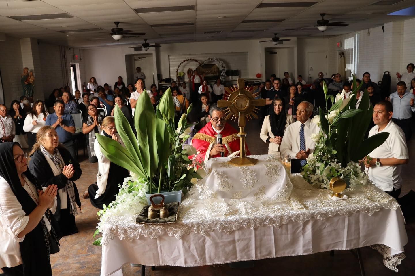 Crowded indoor ceremony with a decorated table, white cloth, candles, and large green floral arrangements