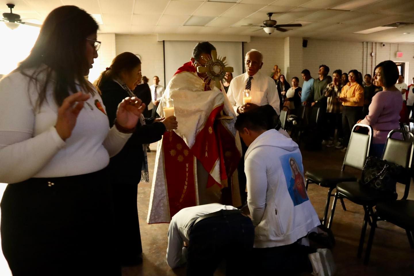 Indoor gathering with people standing in prayer and applause during a ceremony, with a robed officiant at center.