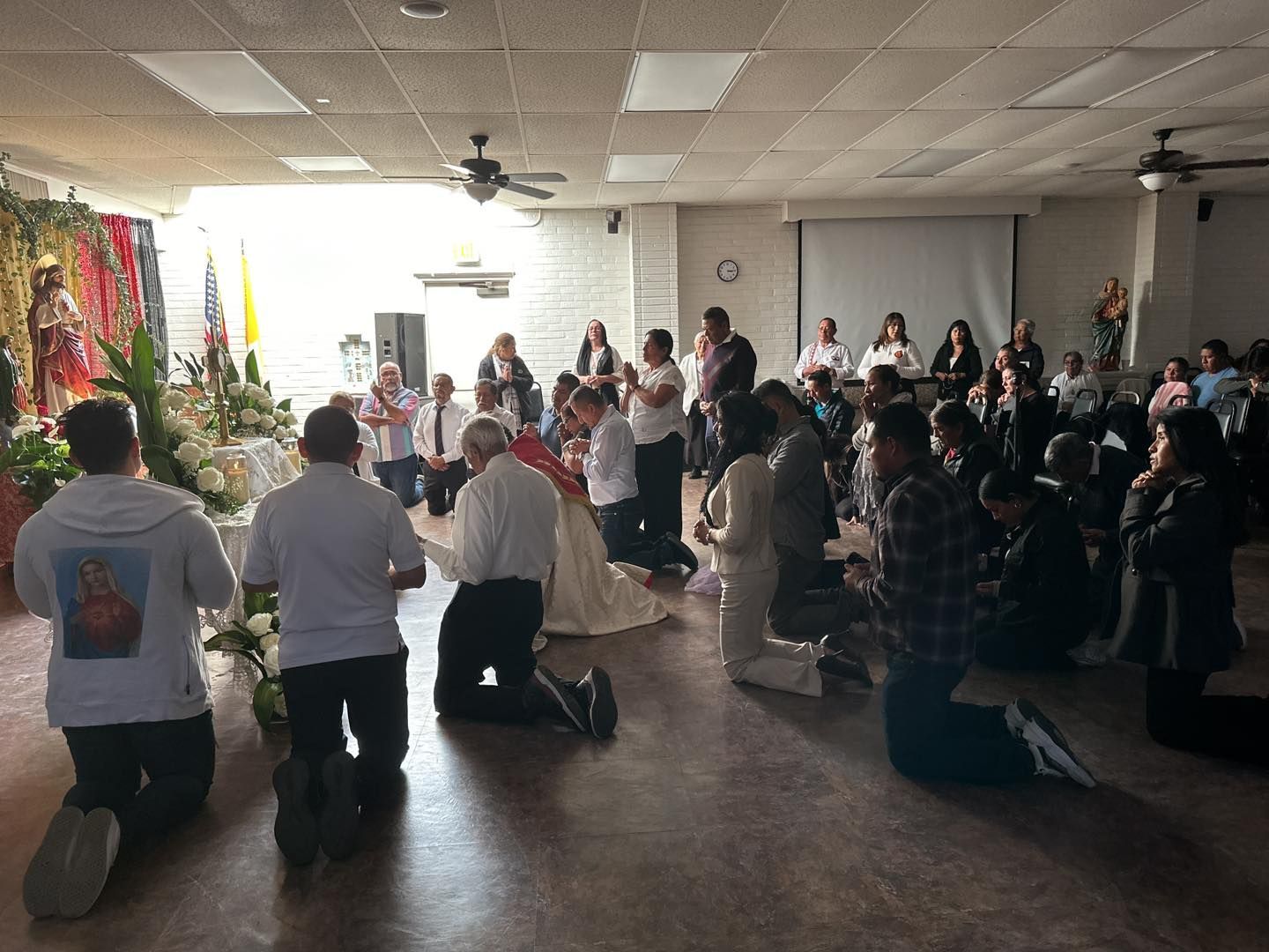 People kneeling in a dim hall during a worship service with a lit Christmas tree and projection screen