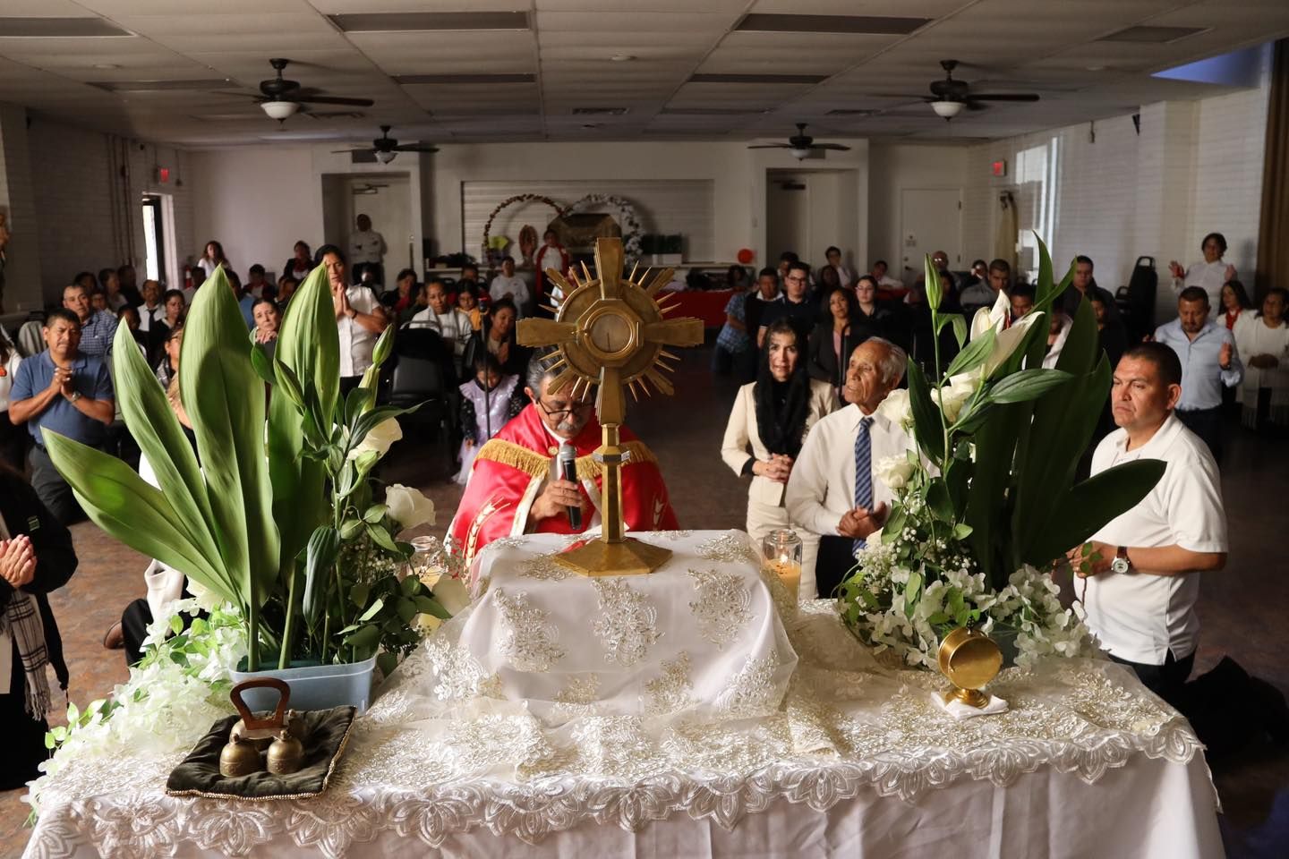 Religious procession inside a hall, with a decorated altar, flowers, and worshippers gathered around it.