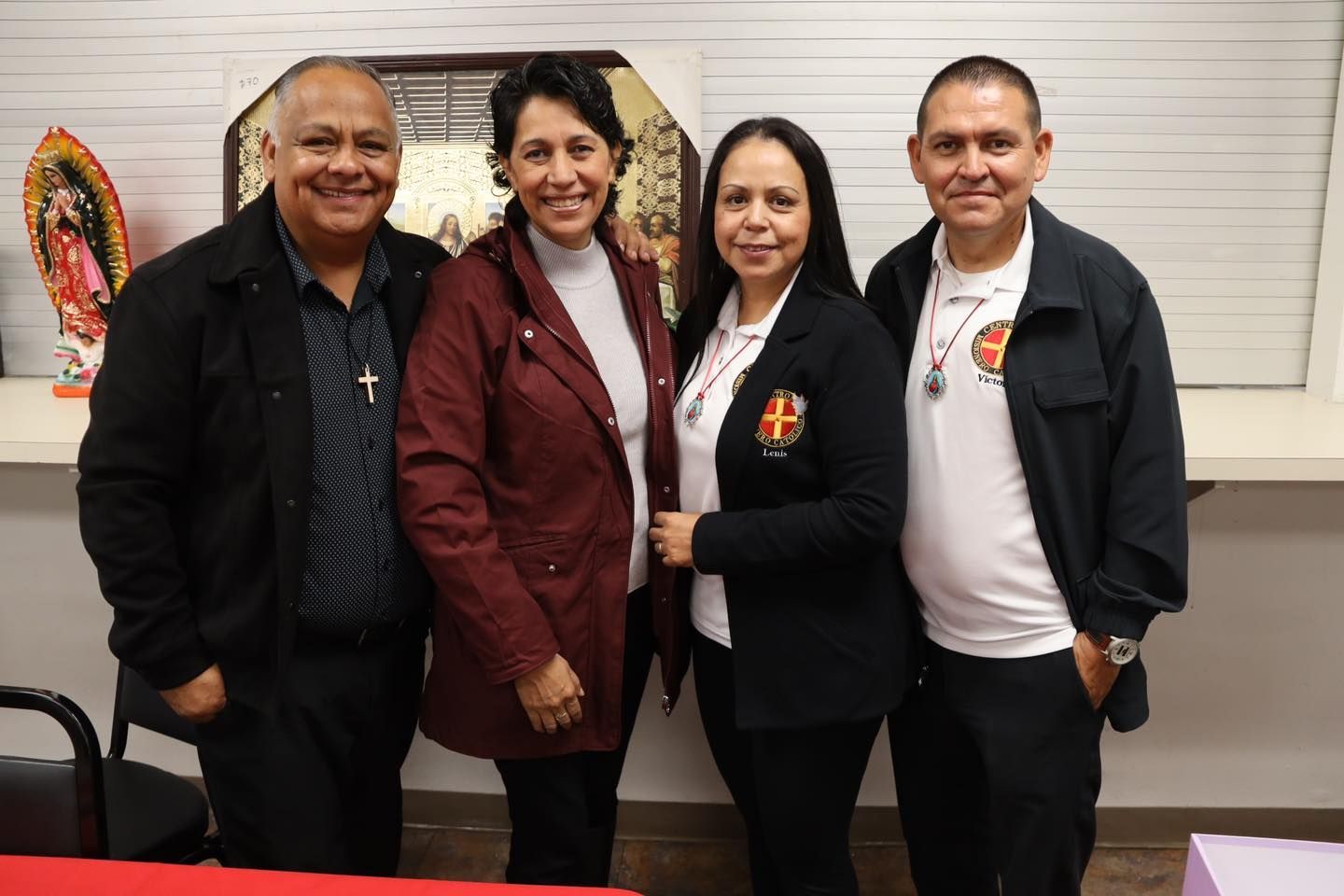 Four people standing together indoors, smiling in front of a window blind.
