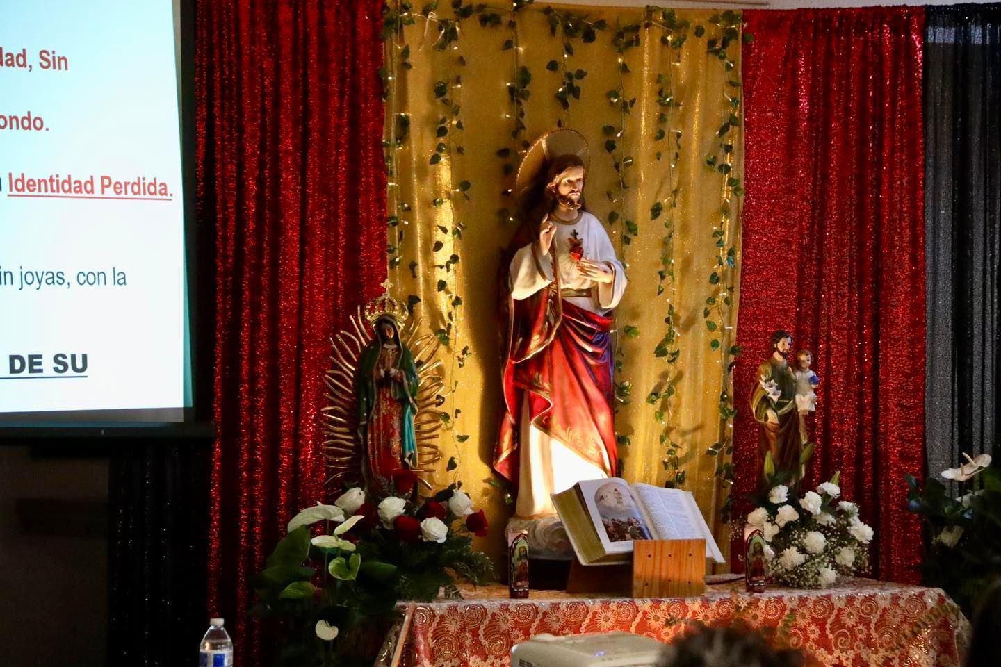 Statue on decorated stage with red and gold curtains and flowers, beside a presentation screen