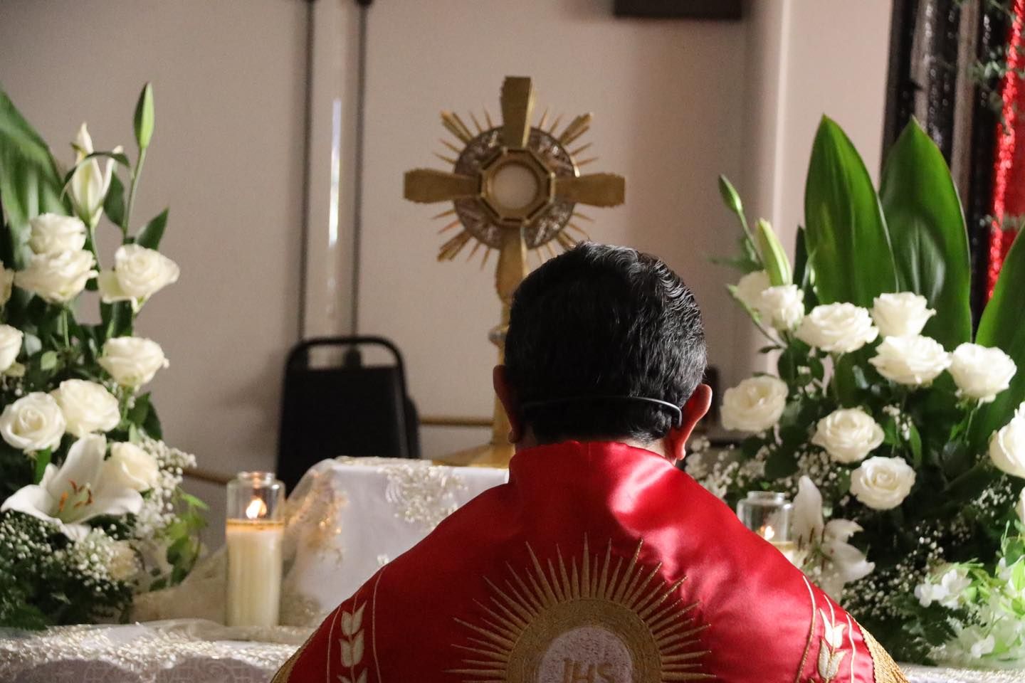 Person in red robe seated at an altar with candles and white flowers, facing a golden religious emblem