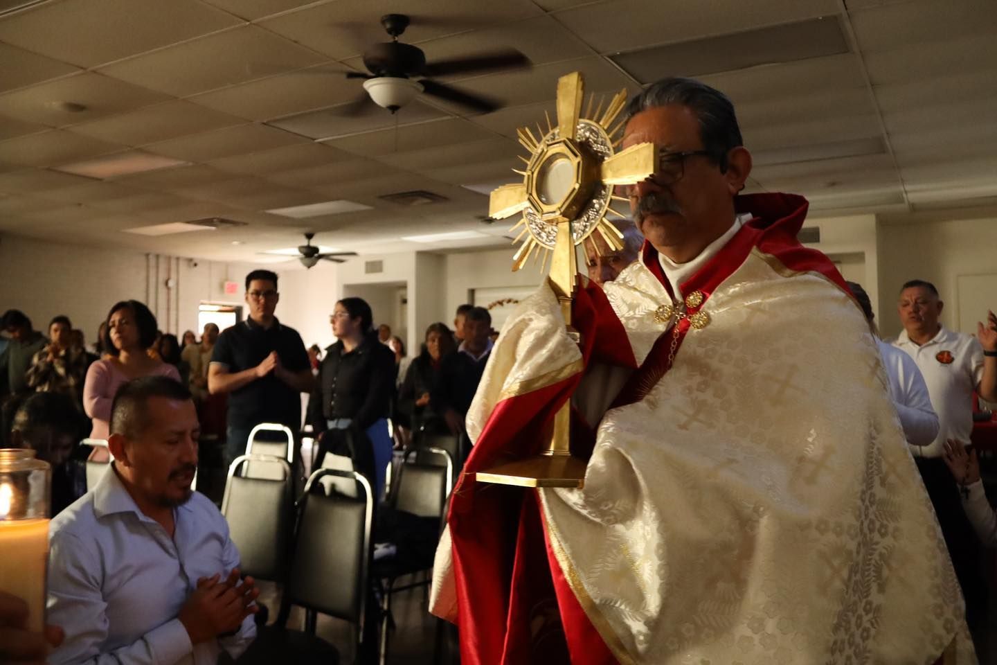 Priest holding a golden monstrance in a church hall, with seated attendees in the background