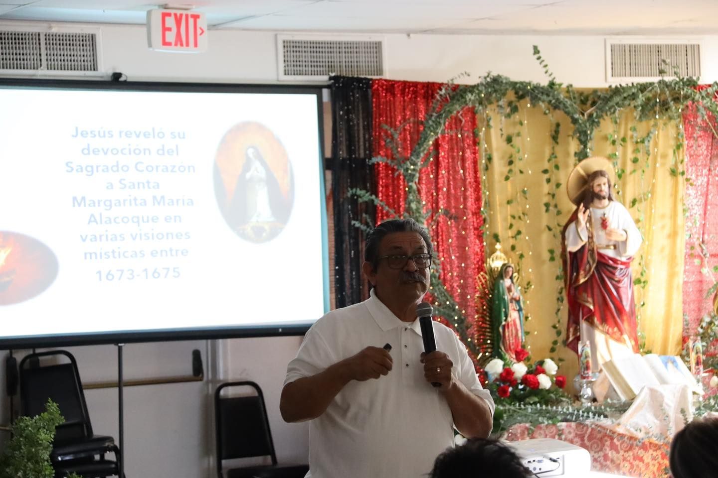 Man speaking into a microphone beside a projection screen and a flower-decorated altar with a statue