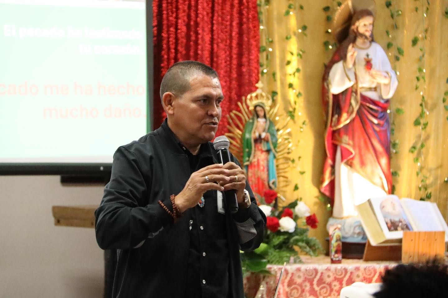 Man speaking into a microphone at a decorated indoor event, with a religious statue and flowers behind him