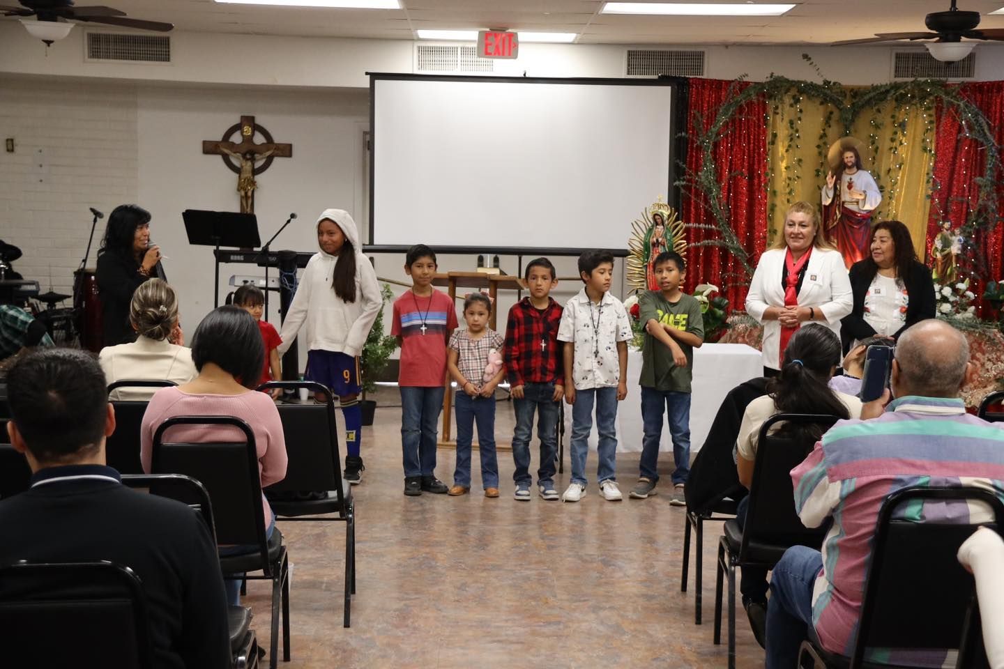 Children stand on a church stage with adults, facing seated attendees during a performance or ceremony.
