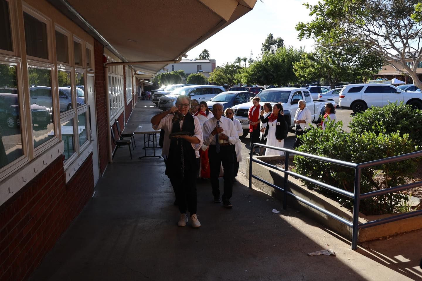People walking along a sunny sidewalk beside parked cars and a building entrance.