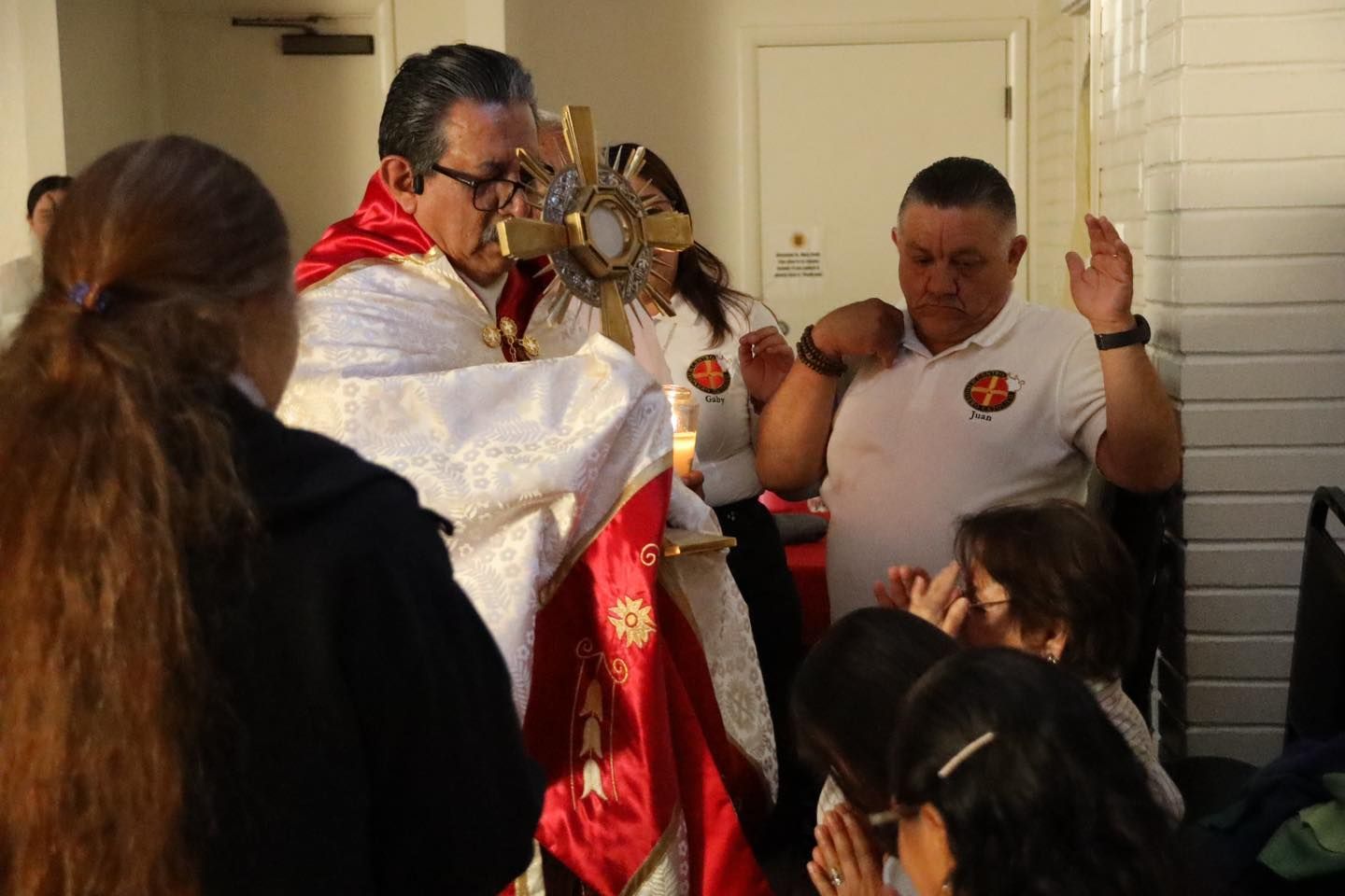 Priest holding a gold cross in a doorway, with people gathered around in a small indoor ceremony