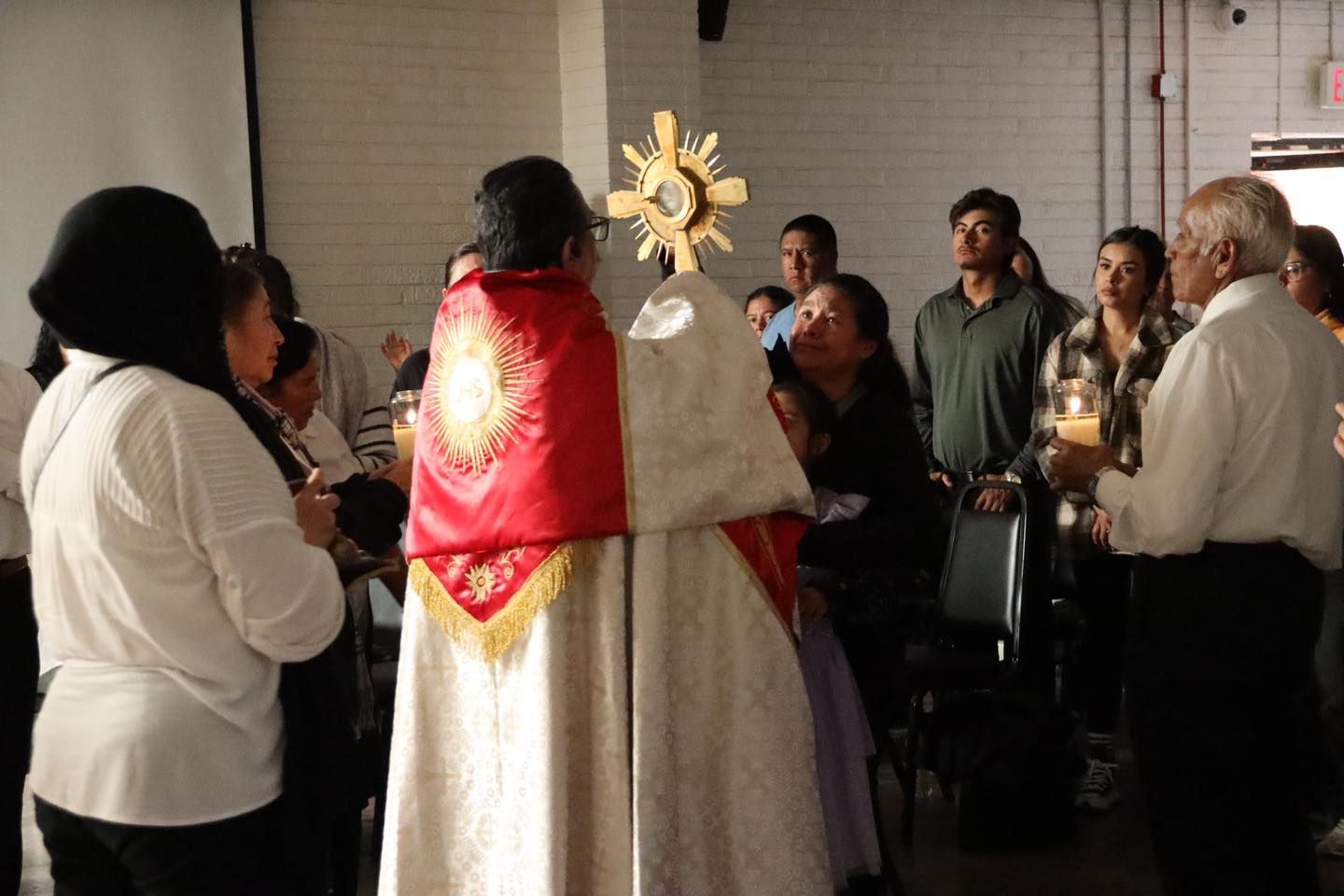Clergy in red and white vestments face a small congregation indoors, with a gold sunburst visible behind them.