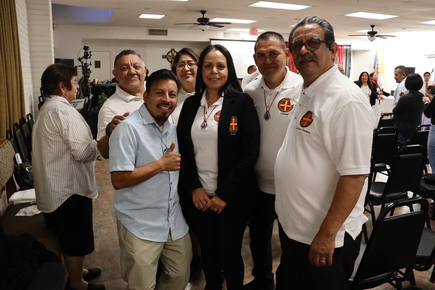 Group of people smiling and posing together at an indoor event, with chairs and attendees in the background.