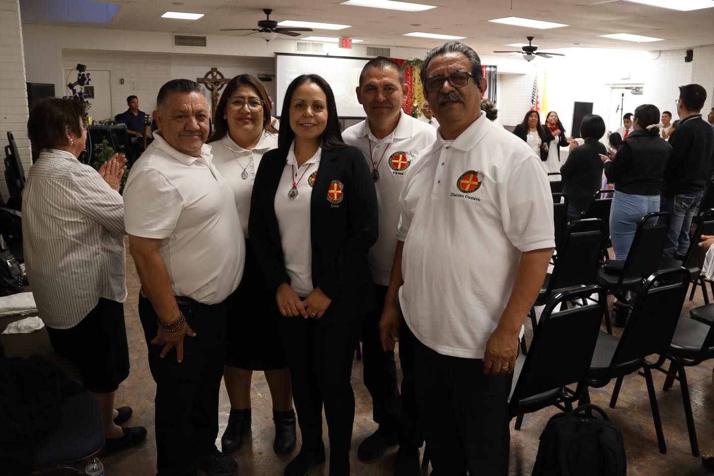 Four people posing indoors at a crowded event, wearing white shirts and black clothing.