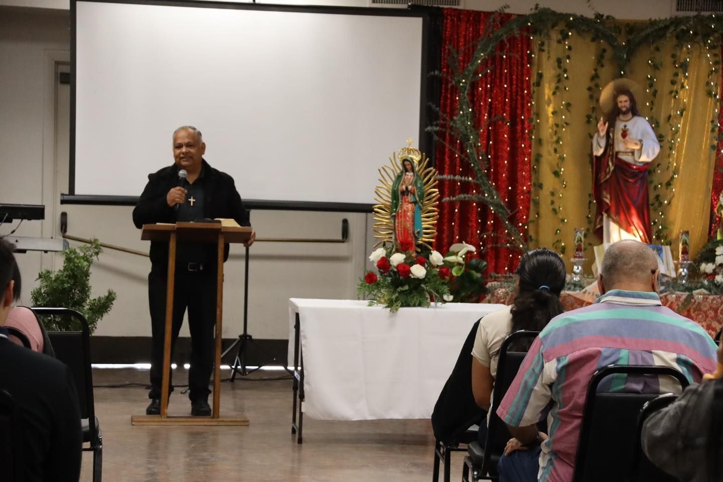Man speaking at a podium in a decorated hall, with seated audience and a projection screen behind him.