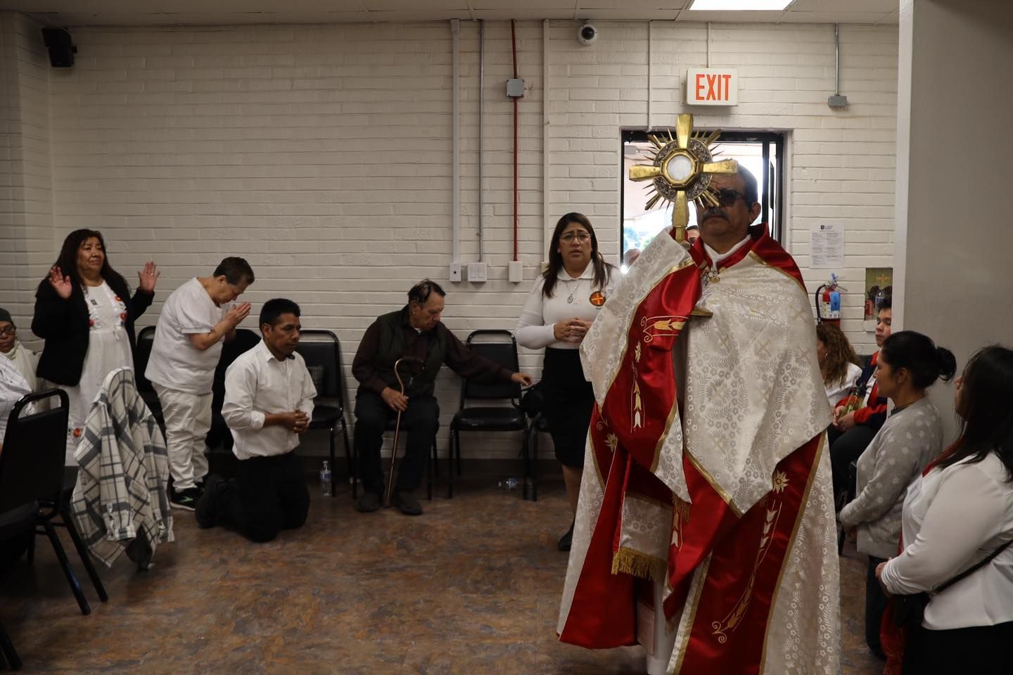 Priest in red and white vestments holding a monstrance during a church service, with parishioners kneeling nearby