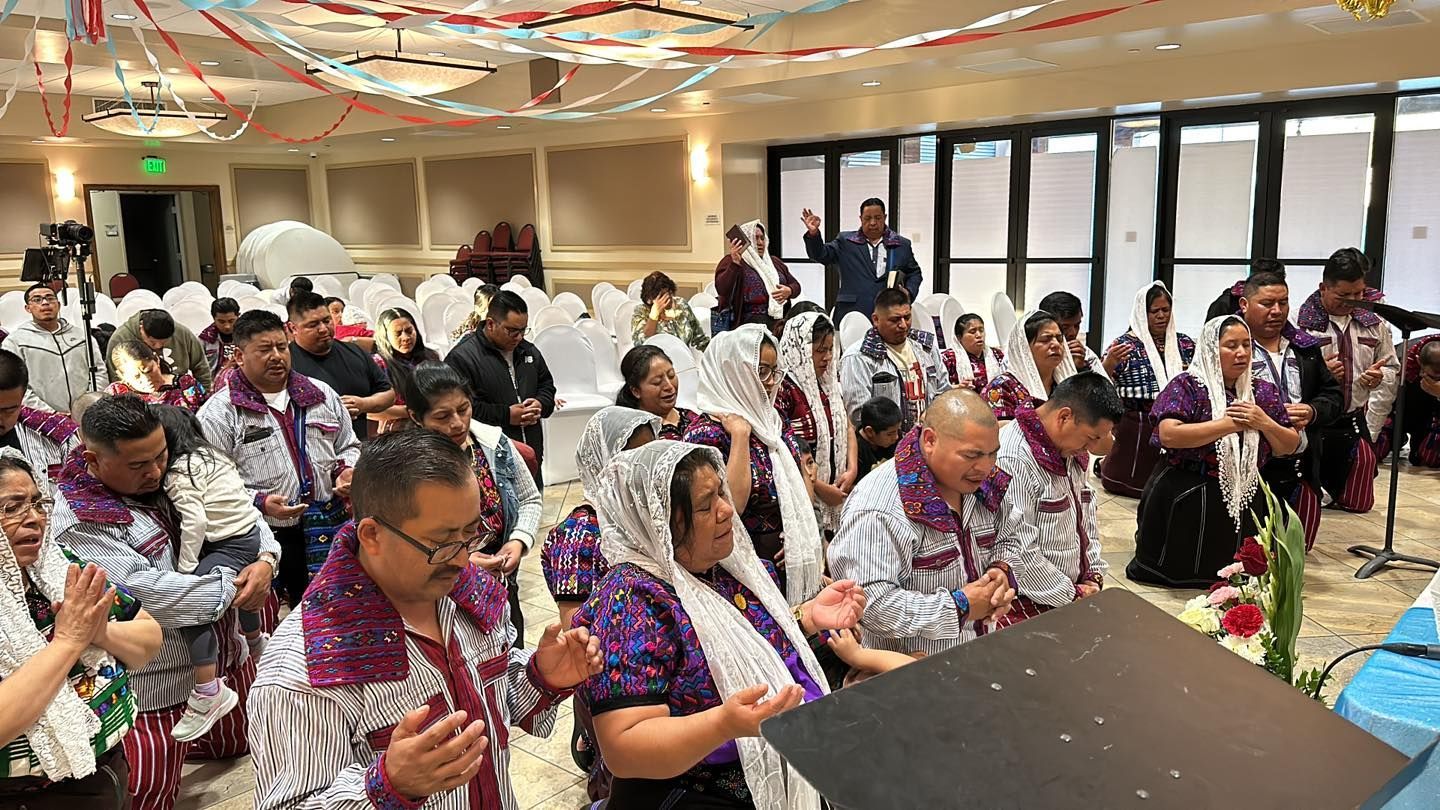 People seated in a decorated hall, many clapping and looking toward a front stage or podium.