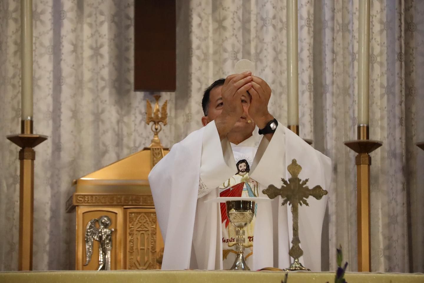 Priest in white vestments stands at altar, covering his face in a church interior.