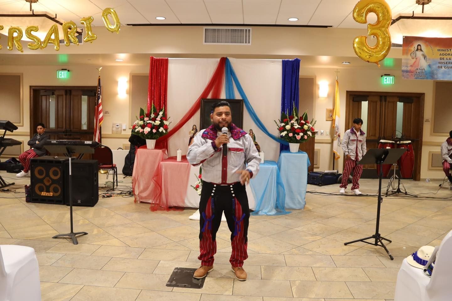 Person speaking into a microphone at a decorated indoor party stage with red, white, and blue drapes.