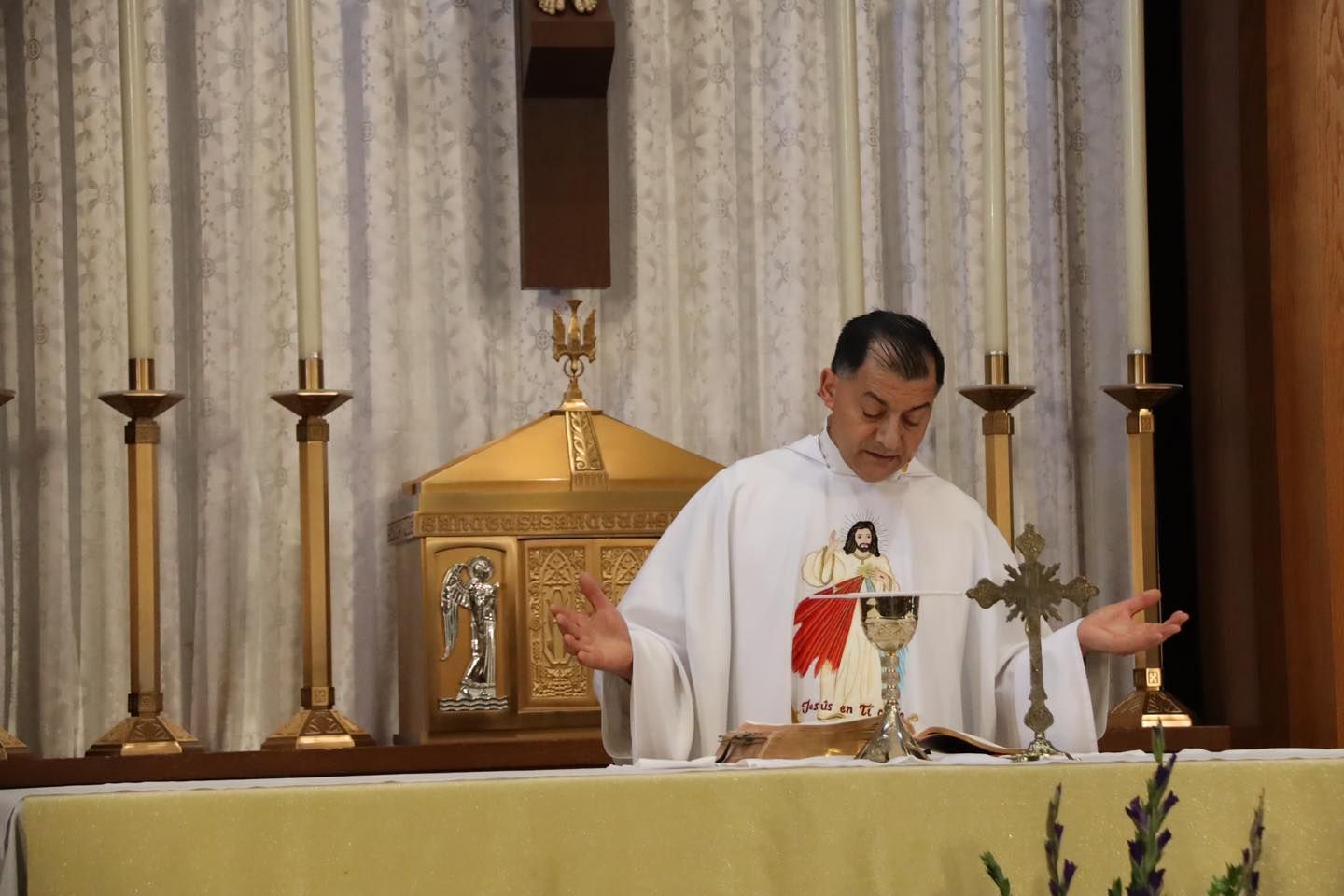Priest in white vestments praying at the altar during Mass, with candles and a tabernacle behind him