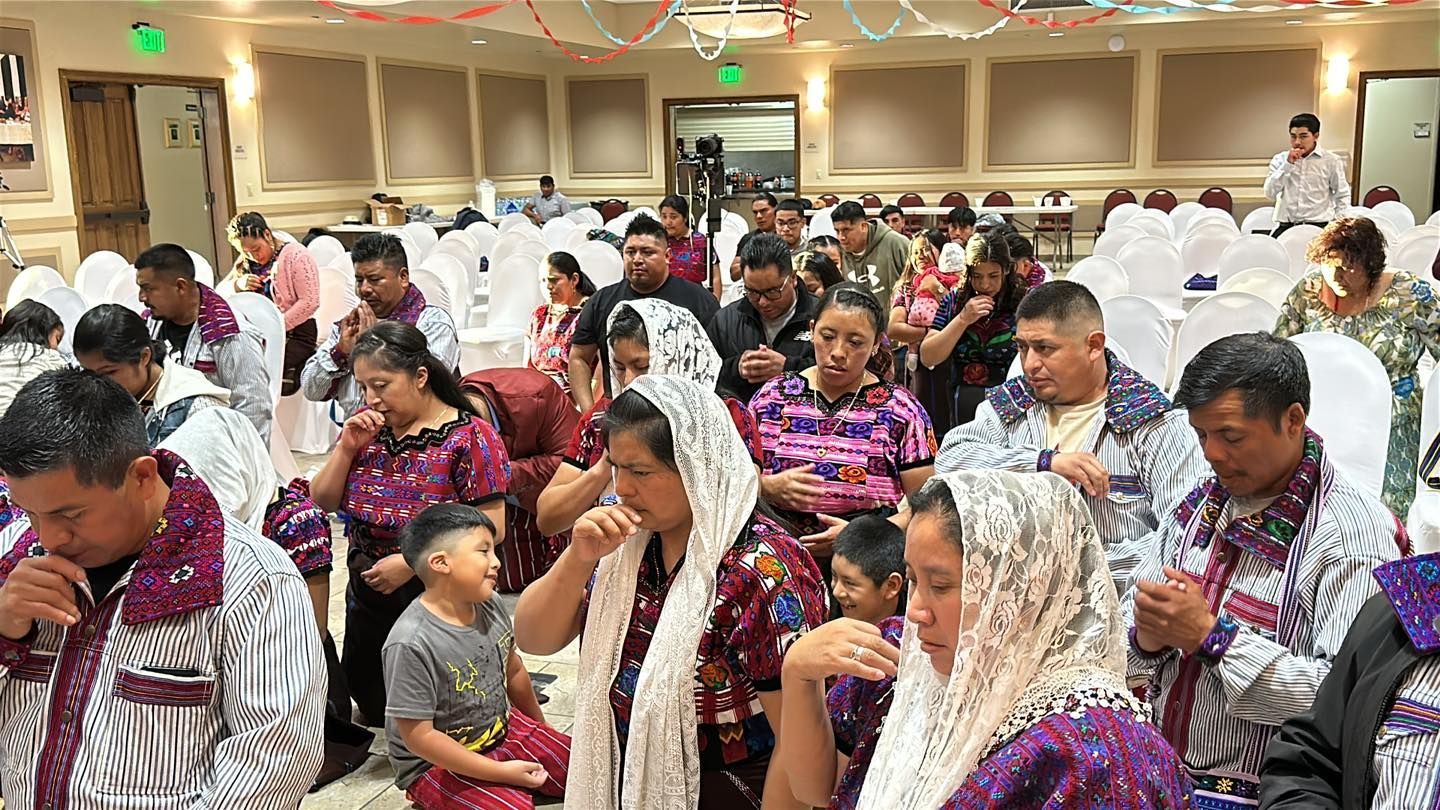 Audience seated in a banquet hall, many wearing purple patterned shawls and listening attentively.