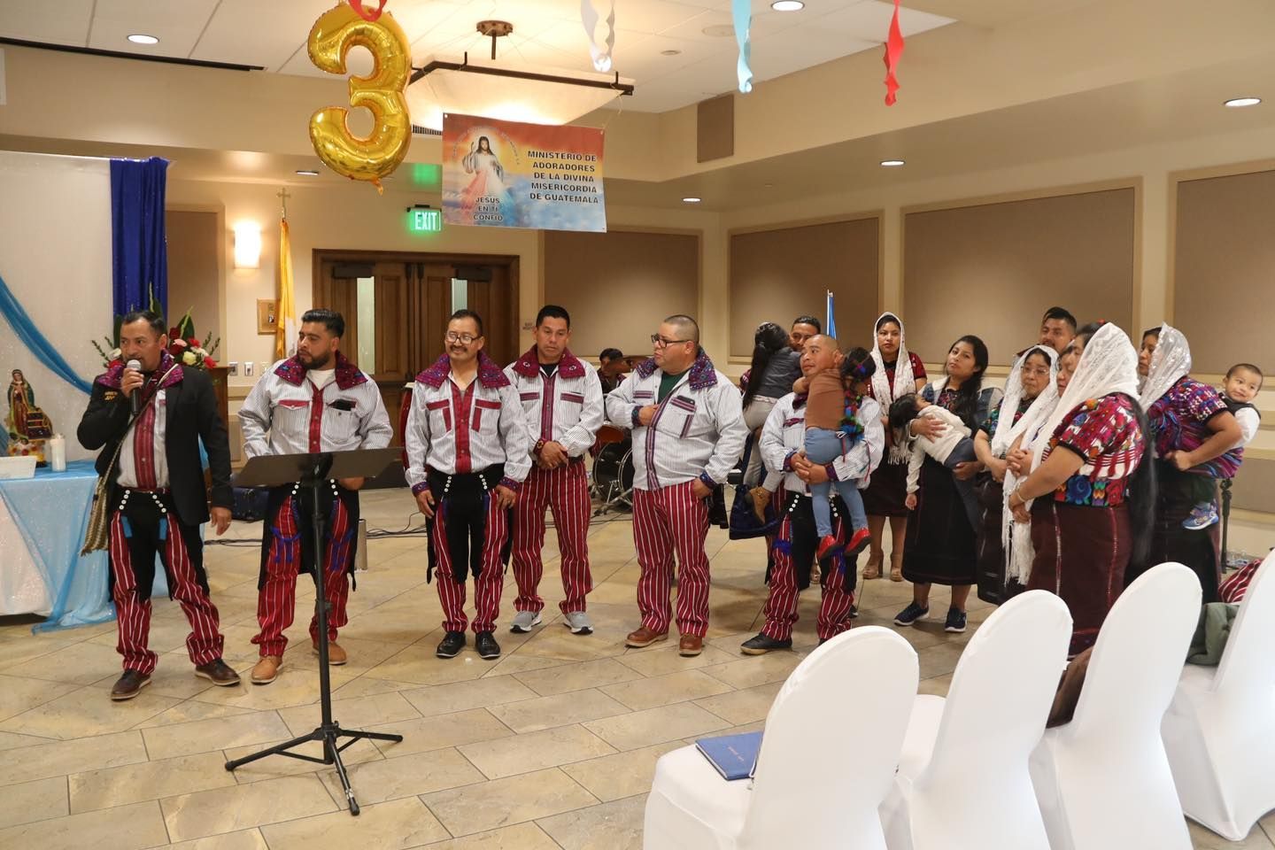 Dancers in red and black costumes perform indoors at a celebration with balloons and a microphone stand.